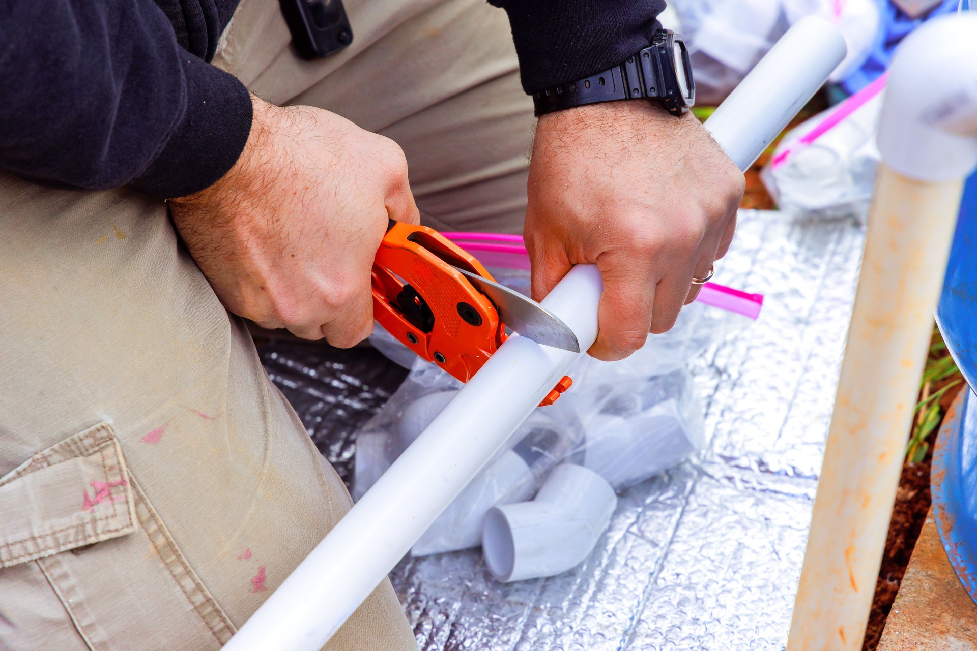 Hands carefully cutting white PVC pipe with a tool during DIY project in outdoor setting near various fittings