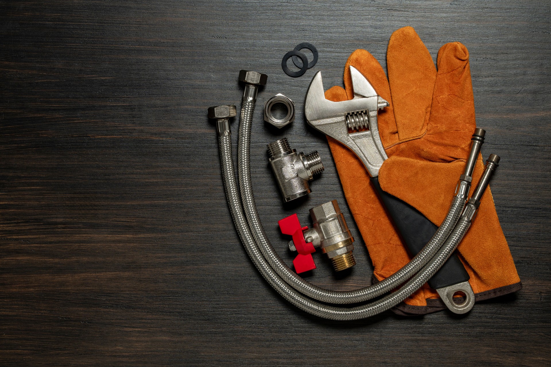 Adjustable wrench, ball valve with red handle, stainless steel tee fittings, leather safety gloves and rubber gaskets on wooden background, top view, copy space, closeup. Plumbing repair concept
